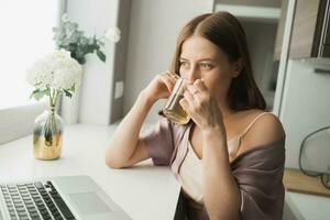 jong vrouw draagt slapen kleren werken Aan laptop computer terwijl zittend Bij leven kamer en drinken thee - sociaal netwerk en werken Bij huis concept foto