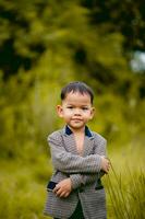 schattig weinig jongen een goed gekleed jongen in een pak in een achtertuin met een gazon en op zoek voor iets interessant. foto