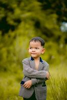 schattig weinig jongen een goed gekleed jongen in een pak in een achtertuin met een gazon en op zoek voor iets interessant. foto