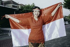 gelukkig glimlachen Indonesisch vrouw vervelend rood kebaya Holding Indonesië's vlag naar vieren Indonesië onafhankelijkheid dag. buitenshuis fotoshoot concept foto