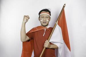 een jong Aziatisch Mens met een gelukkig geslaagd uitdrukking vervelend rood top en hoofdband terwijl Holding Indonesië's vlag, geïsoleerd door wit achtergrond. Indonesië's onafhankelijkheid dag concept. foto