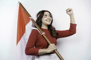 een jong Aziatisch vrouw met een gelukkig geslaagd uitdrukking vervelend rood top en hoofdband terwijl Holding Indonesië's vlag, geïsoleerd door wit achtergrond. Indonesië's onafhankelijkheid dag concept. foto