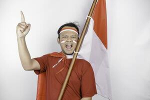 een verlichten jong Aziatisch Mens is vervelend rood t-shirt en Holding Indonesië's vlag looks geïnspireerd door ideeën, geïsoleerd door wit achtergrond foto