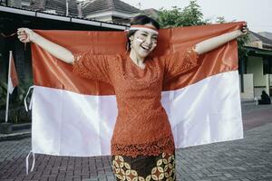 gelukkig glimlachen Indonesisch vrouw vervelend rood kebaya Holding Indonesië's vlag naar vieren Indonesië onafhankelijkheid dag. buitenshuis fotoshoot concept foto