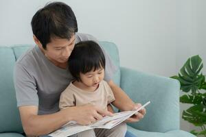 gelukkig Aziatisch vader kom tot rust en lezen boek met baby tijd samen Bij huis. ouder zitten Aan sofa met dochter en lezing een verhaal. leren ontwikkeling, kinderopvang, lachend, opleiding, verhaal vertellen, oefening. foto