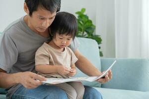 gelukkig Aziatisch vader kom tot rust en lezen boek met baby tijd samen Bij huis. ouder zitten Aan sofa met dochter en lezing een verhaal. leren ontwikkeling, kinderopvang, lachend, opleiding, verhaal vertellen, oefening. foto