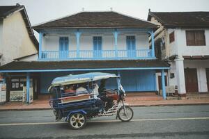 luangprabang lao-juli20,2023 , drie wiel motorfiets voorbijgaan mooi oud gebouw in hart van luangprabang stad- , luang prabang is een van wereld erfgoed plaats van UNESCO foto