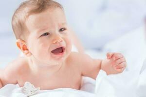 portret van schattig kleuter jongen aan het liegen Aan bed. pasgeboren kind in slaapkamer. baby leugens Aan zijn buik verhoogt de hoofd en huilen foto