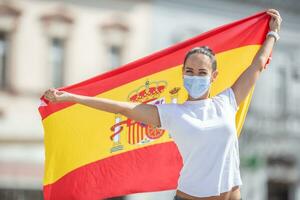 glimlachen meisje houdt een Spaans vlag achter haar terug vervelend een gezicht masker foto
