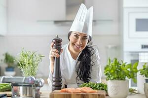 vrouw chef in toque blancheren, wit hoed, en uniform houdt een peper Slijper gedurende vis en veg maaltijd voorbereiding foto