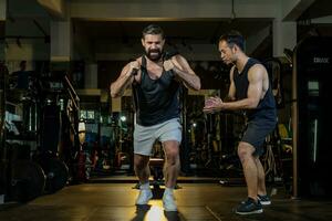 Kaukasisch Mens met sterk en gespierd lichaam opleiding Aan kern gewicht opleiding door touw trekken met zijn ondersteunen trainer bemoedigend binnen Sportschool voor vooruitgaan oefening en training foto