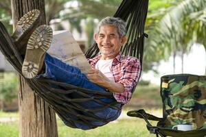 gelukkig senior mannetje met grijs haar- en baard draagt geruit overhemd kom tot rust Bij toevlucht tuin Aan zomer, volwassen Mens houdende Aan touw hangmat, glimlach en kijken Bij een papier kaart, begrip van foto