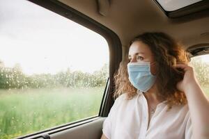 elegant jong Carly vrouw in medisch masker op zoek uit venster terwijl zittend Aan terug stoel van auto Aan wazig achtergrond met zonsondergang foto