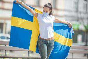 brunette staand buitenshuis looks Bij de Zweeds vlag ze houdt vast, met een gezicht masker Aan foto