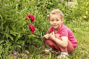 een klein meisje zit glimlachend bij een bloembed in de tuin van roze pioenrozen