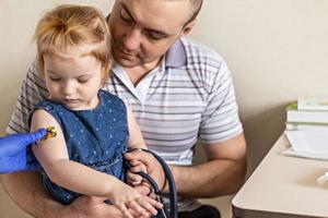 vaccinatie van een klein meisje in de armen van haar vader in de spreekkamer van het coronavirus foto