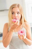 jong blond meisje eet roze donuts in huis keuken met smaak emoties foto