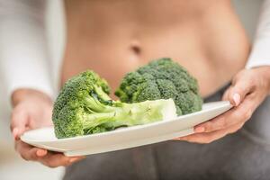 jong vrouw met sport- figuur in de keuken Holding in haar handen en een bord van broccoli foto