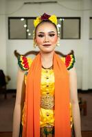 een traditioneel Indonesisch danser staand in een geel kostuum en een oranje sjaal hangende naar beneden haar lichaam foto