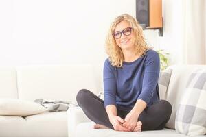 vrouw met bril zit barefood Aan een sofa in een licht leven kamer, glimlachen Bij de camera foto