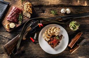 rustiek houten tafel met een rauw hert hertenvlees, heerlijk eigengemaakt knoedel en rozemarijn. tussen de bord vol van hertevlees goulash en knoedel leugens een jacht- geweer met kogels en een mes foto