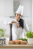 professioneel vrouw chef vol vertrouwen leunt Aan peper Slijper met ingrediënten voor Koken in voorkant van haar foto