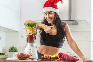 mooi fit vrouw in Kerstmis hoed leunt tegen de blender vol van gezond fruit en veg in een blender foto