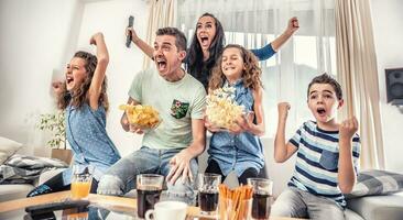 familie aan het kijken sport- bij elkaar passen Aan TV Bij huis, juichen en geschreeuw doel met handen omhoog, morsen chips en popcorn van opwinding foto