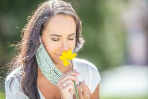 mooi vrouw geniet de geur van een bloem met voor de helft van haar gezicht masker uit, hebben Gesloten ogen Aan een zonnig dag in de natuur foto