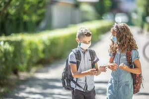 twee jong vrienden klasgenoten met gezicht maskers praten Aan hun manier naar school- gedurende de covid-19 quarantaine foto