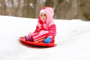 schattig weinig meisje glijden naar beneden van de heuvel top gedekt met wit en pluizig sneeuw. een van de kinderen favoriete werkzaamheid in winter tijd. foto