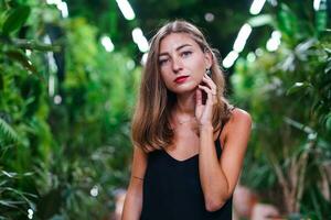 dichtbij omhoog mooi vrouw portret tussen tropisch groenen. jong vrouw vervelend zwart jurk Aan dun riemen staat tussen groen planten binnen ogangery. meisje met lang haar- poseren voor camera. foto