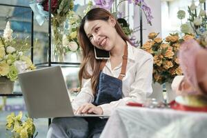 een jong Aziatisch vrouw bloemist baasje, werken met laptop, verkoop bloemen afspreken, pratend Aan mobiel telefoon in kleurrijk bloem winkel op te slaan met een mooi bundel van bloesems, en e-commerce bedrijf. foto