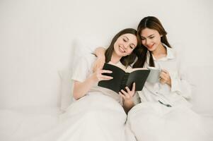aantrekkelijk lesbienne paar gebruik mobiel telefoon, tablet kijk maar film Aan bed. mooi broer of zus in pyjama aan het liegen naar beneden met deken in slaapkamer samen, in ochtend- foto