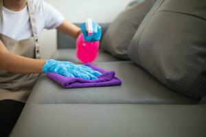 schoonmaak personeel is afvegen kleding met schoonmaakster en ontsmettingsmiddel Aan de oppervlakte van sofa naar maken de sofa schoon met schoonmaak producten en vrij van kiemen vastklampen naar oppervlakte van de sofa in leven kamer. foto