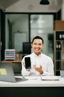 zelfverzekerd Aziatisch Mens met een glimlach staand Holding kladblok en tablet Bij de kantoor. foto