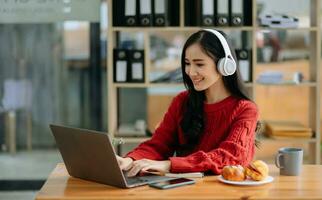 aantrekkelijk gelukkig jong Aziatisch leerling aan het studeren Bij de college bibliotheek, zittend Bij bureau, gebruik makend van een laptop computer, tablet en hoofdtelefoons hebben een video chatten. foto