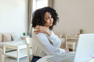 portret van jong benadrukt vrouw zittend Bij huis kantoor bureau in voorkant van laptop, aanraken pijn doen terug met gepijnigd uitdrukking, lijden van rugpijn na werken Aan pc foto