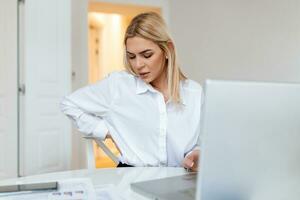 portret van jong benadrukt vrouw zittend Bij huis kantoor bureau in voorkant van laptop, aanraken pijn doen terug met gepijnigd uitdrukking, lijden van rugpijn na werken Aan pc foto