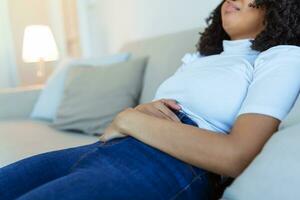 Afrikaanse Amerikaans vrouw in pijnlijk uitdrukking Holding handen tegen buik lijden menstruatie- periode pijn, aan het liegen verdrietig Aan huis bed, hebben buik kramp in vrouw Gezondheid concept foto