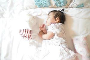 schattig slaperig 1 jaar oud Aziatisch babymeisje dat op zacht beddengoed slaapt met een pop, pasgeboren concept.