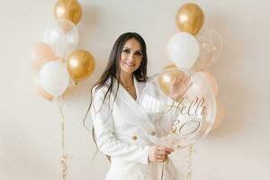 positief vrouw vieren verjaardag gekleed in elegant veer jurk poseren met verjaardag ballon in de omgeving van opgeblazen ballonnen Bij huis foto