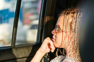 een nadenkend tiener meisje met afro-vlechten en zonnebril looks uit de venster terwijl zittend in de auto. weg reis foto