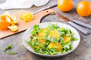 eetpatroon vegetarisch vitamine salade van oranje plakjes en een mengen van rucola, snijbiet en mizun bladeren Aan een bord en een snijdend bord met geschild oranje Aan de tafel foto