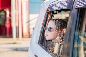 een schattig tiener- meisje in zonnebril met afro-vlechten looks uit de venster terwijl zittend in de auto. weg reis foto