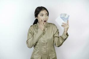 een geschokt jong regering arbeider vrouw is vervelend khaki uniform Holding contant geld geld in Indonesisch roepia geïsoleerd door wit achtergrond foto