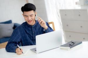 jonge aziatische man praten over telefoon en werken vanuit huis met laptopcomputer, freelance man schrijven op notebook en spreken op smartphone, thuis blijven, huiselijk leven, zaken- en communicatieconcept. foto
