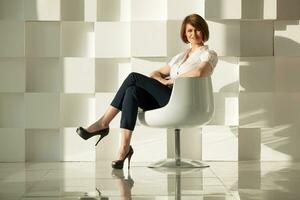elegant volwassen vrouw zittend in stoel tegen van wit modern muur foto