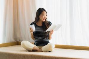 portret mooie jonge aziatische vrouw die boek leest en koffiekopje of mok vasthoudt op de bank in de woonkamer foto