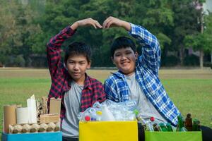 Aziatisch jongen in plaid overhemd zittend Aan speelplaats van school- met divers dozen van gescheiden kledingstukken of afval binnen, concept voor eco vrienden en globaal opwarming campagne, zacht en selectief focus. foto